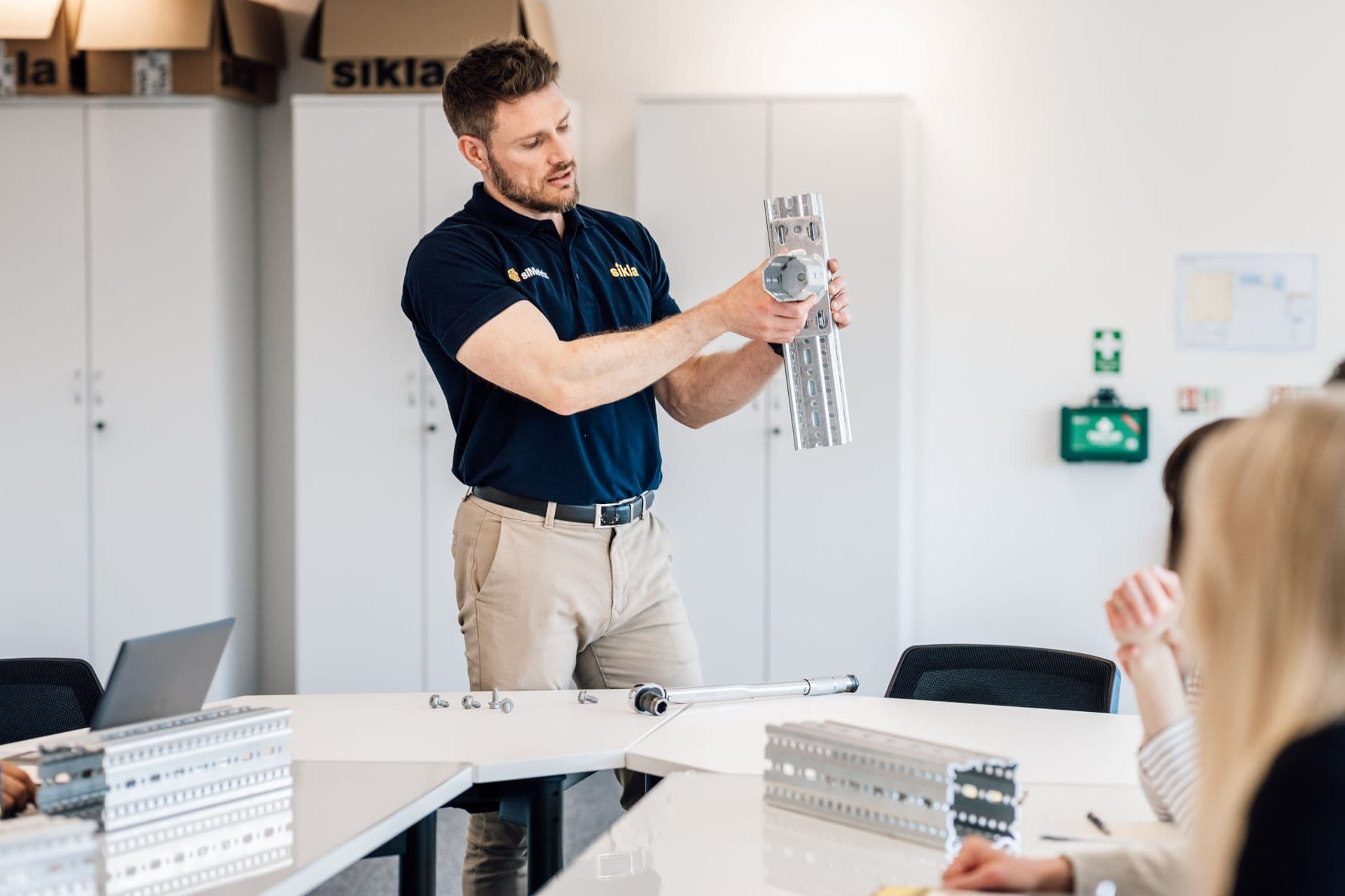 Man in navy shirt demonstrating a metal bracket and bolts to colleagues around a table.