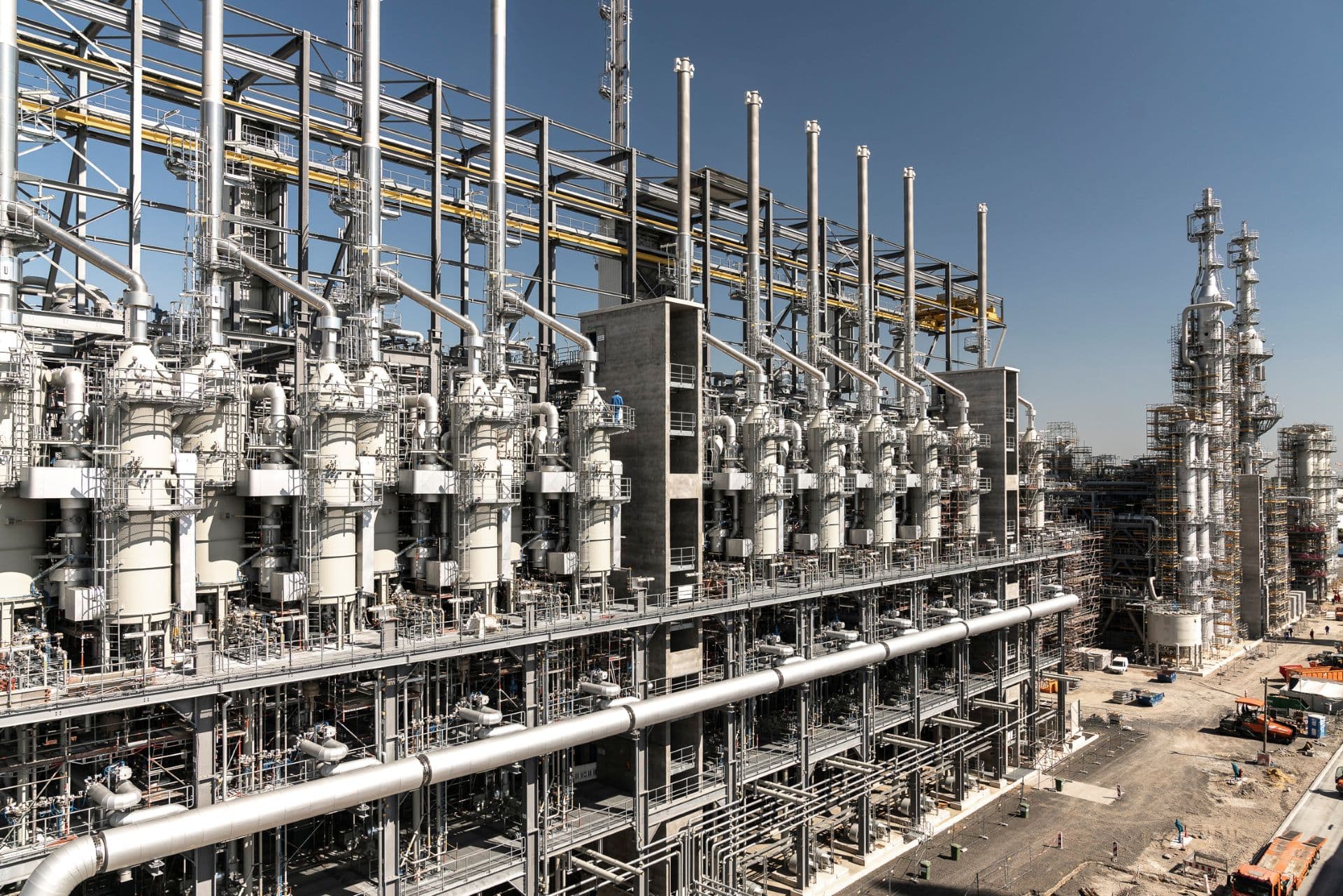 Industrial plant with rows of tall process columns, networks of pipes, walkways and scaffolding under a clear sky