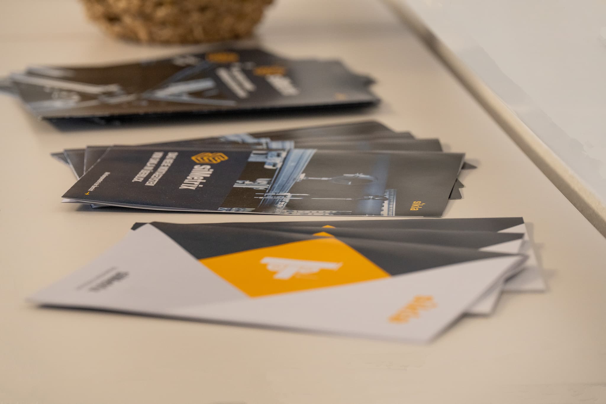 Stacked promotional brochures on a table, foreground white-and-yellow leaflet with a bell icon and darker brochures blurred in the background