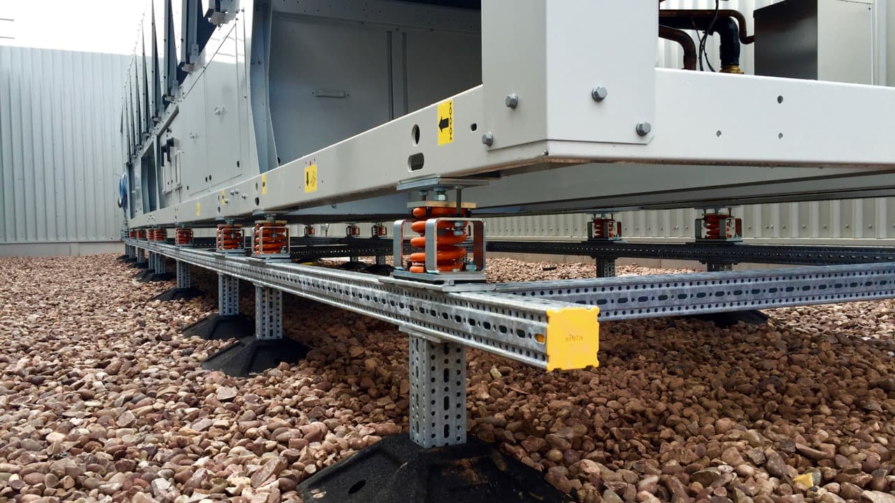 Close-up view of industrial equipment on vibration isolators and metal steel beams, placed on gravel ground near a metal building wall.