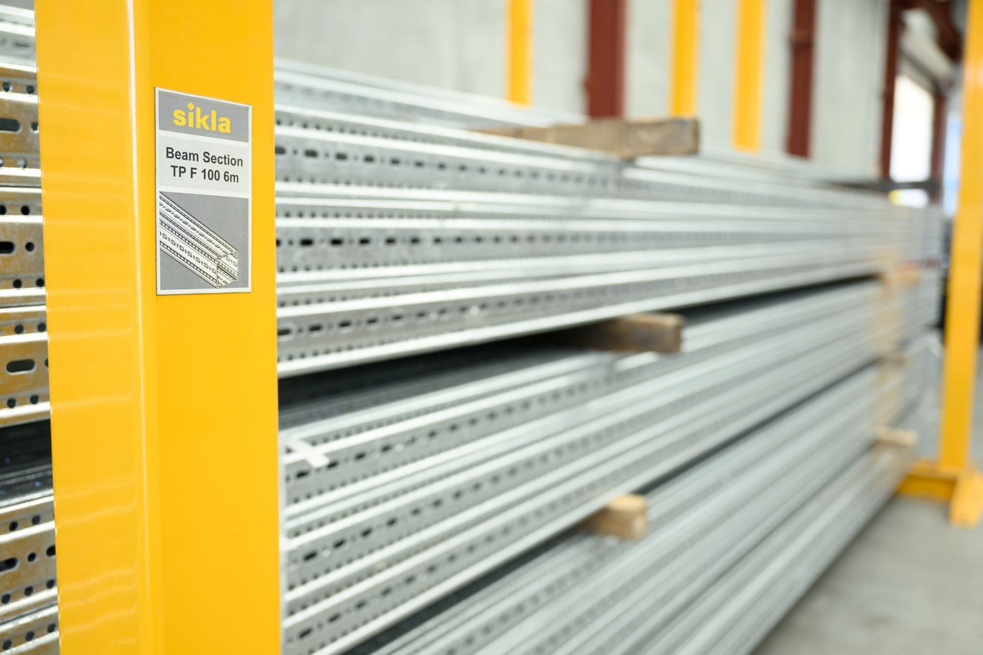 Stacked steel beams on yellow shelving in a warehouse, with a label reading "sikla Beam Section TP F 100 6m" on the left.
