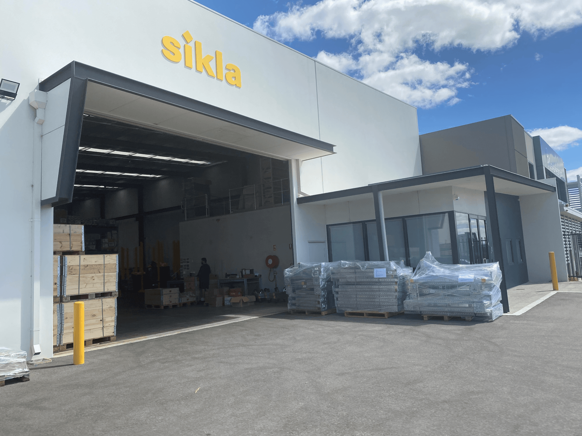 Warehouse exterior with a large open garage door, pallets of goods outside, and "Sikla" sign on the building