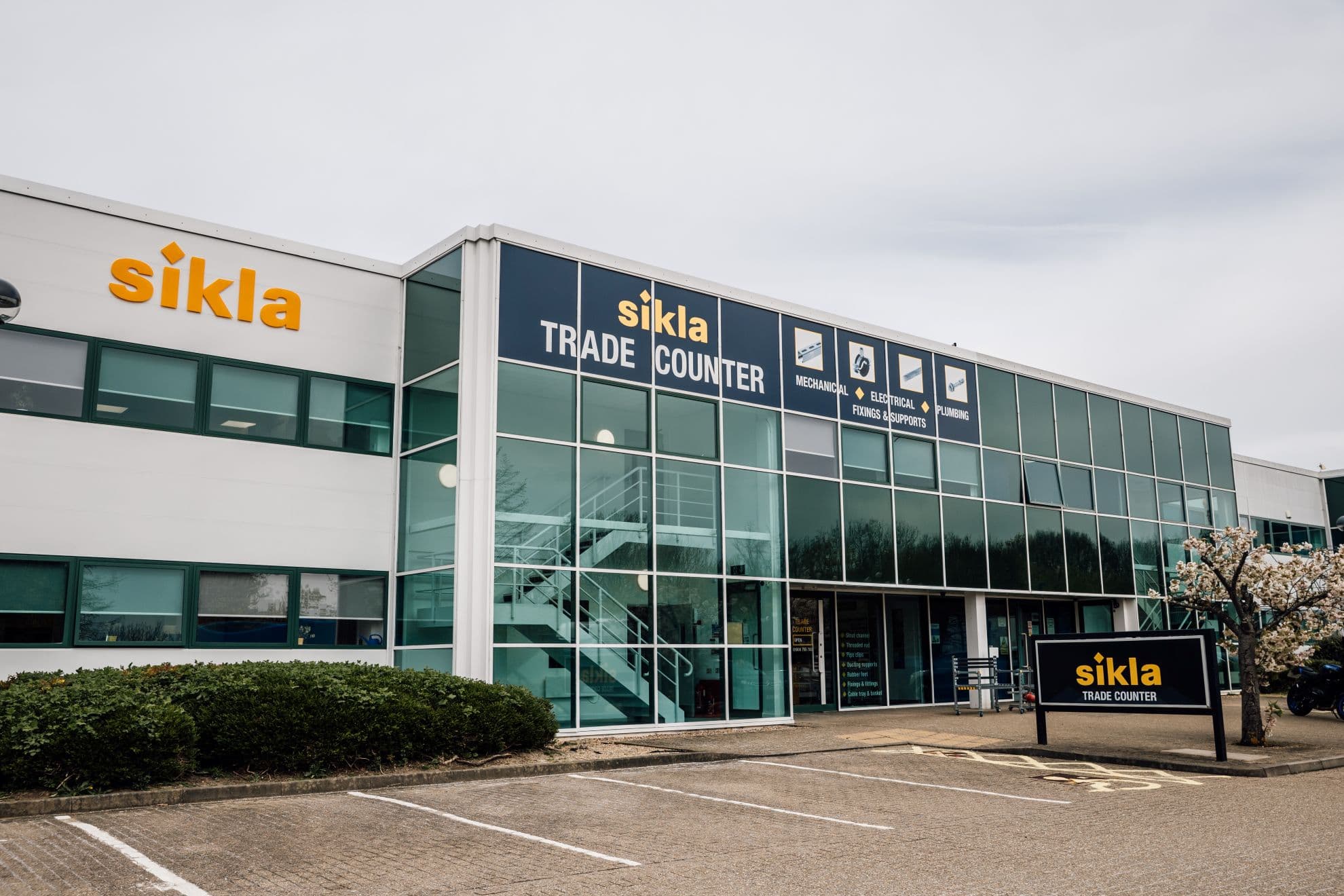 Modern building with large glass windows and signage for Sikla Trade Counter. Bushes and a tree in the foreground.